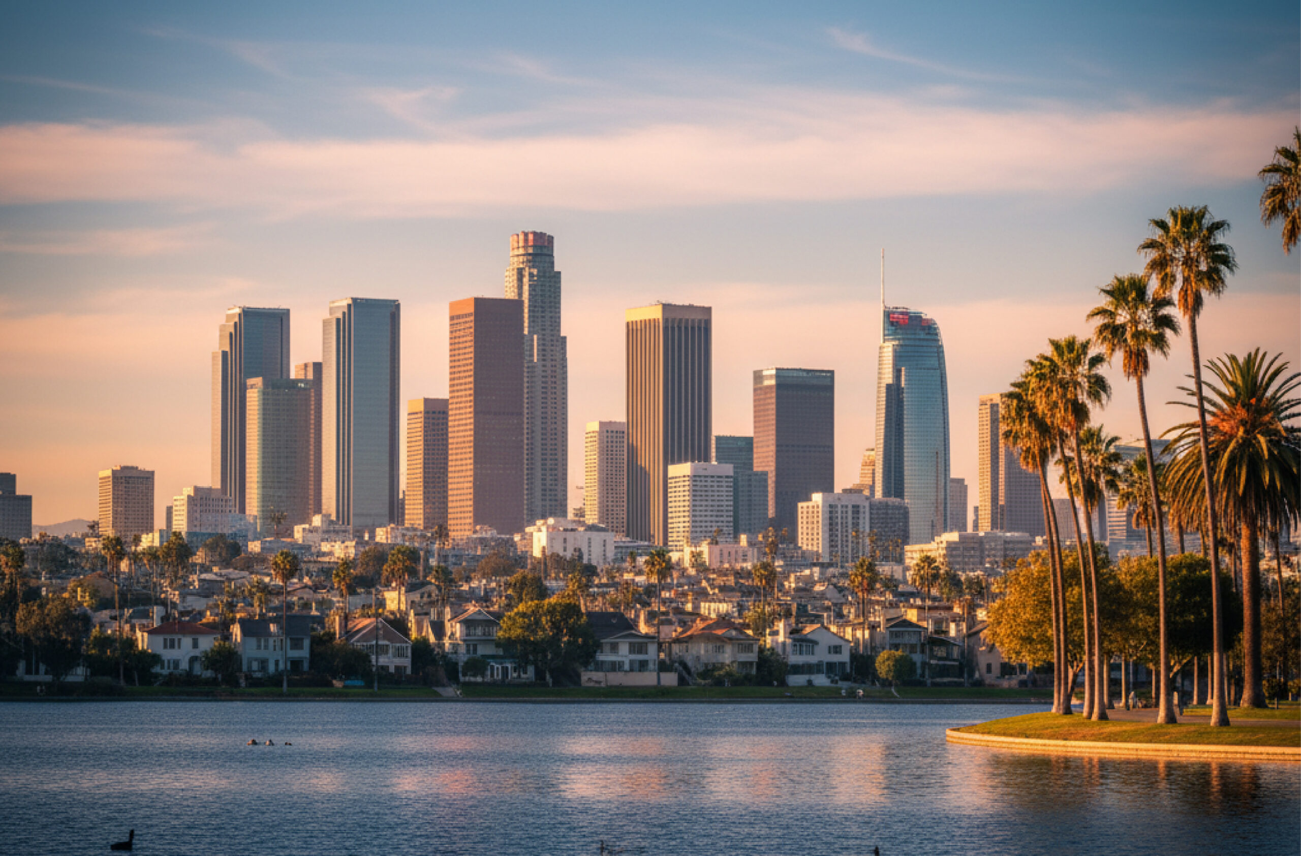 Los Angeles skyline at sunset with palm trees and a lakeside foreground.