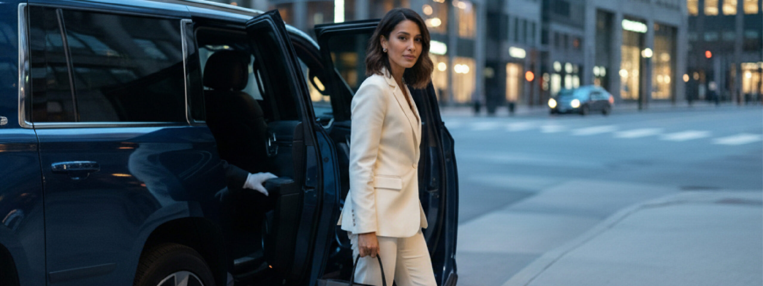 Woman in a cream suit steps from a blue SUV while a gloved chauffeur holds the door on a city street at dusk.