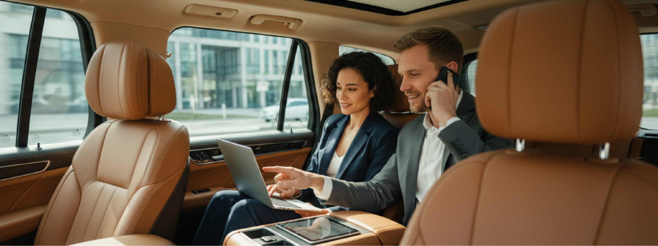 Two business passengers sit in the rear of a luxury SUV; one talks on the phone while the other works on a laptop.
