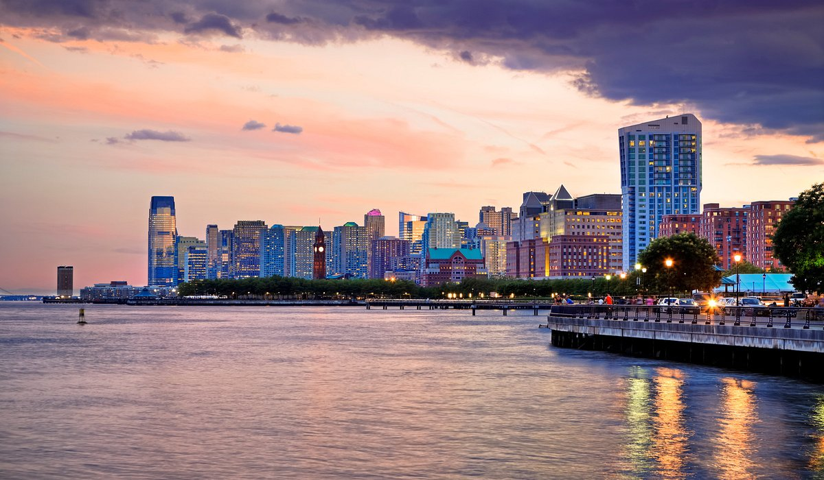 Waterfront skyline at sunset with a riverside promenade and city buildings reflected on the water.