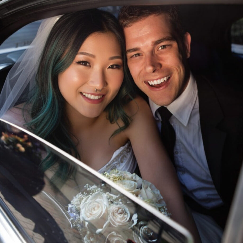 Smiling newlyweds sit together in the back of a car, bride holding a white rose bouquet. Jiberia Limo Service Wedding Transportation