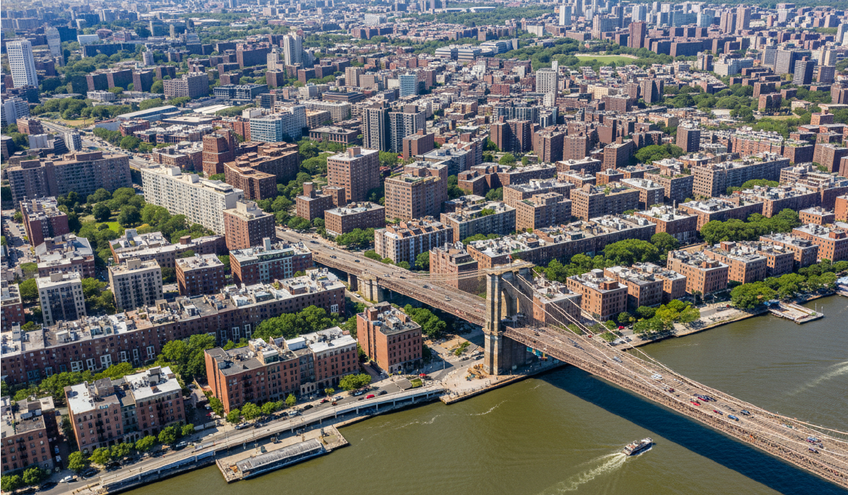 Aerial view of the Brooklyn Bridge spanning the East River toward Manhattan, with surrounding residential blocks and traffic visible.